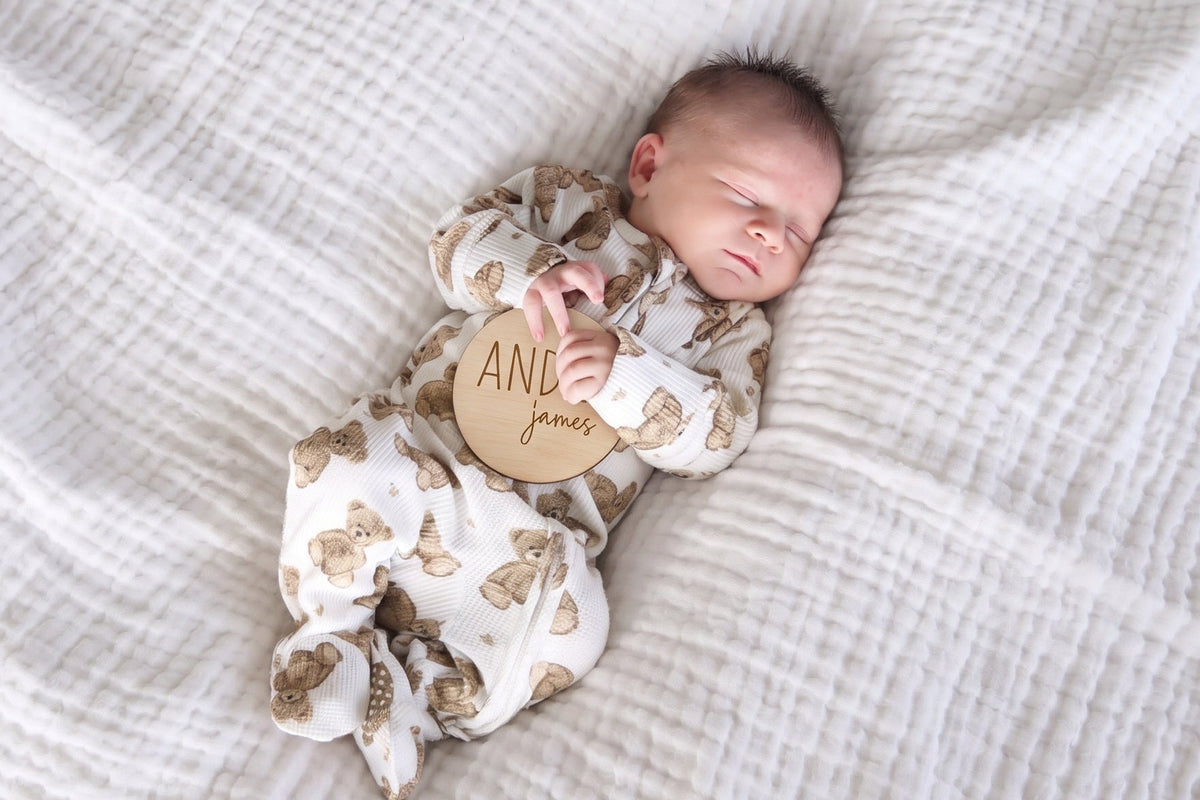 A sleeping newborn baby wearing a Caden Lane bear footed onesie, lying on a white textured muslin blanket. The baby is holding a round wooden tag engraved with 'AND JAMES'.