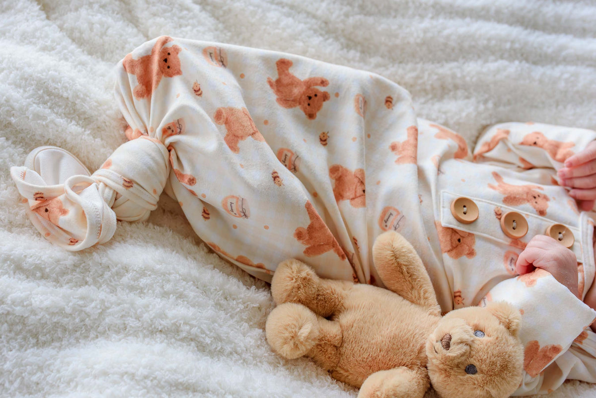 Newborn wearing a Caden Lane honey bear print knotted gown, lying on a fluffy white blanket next to a plush teddy bear