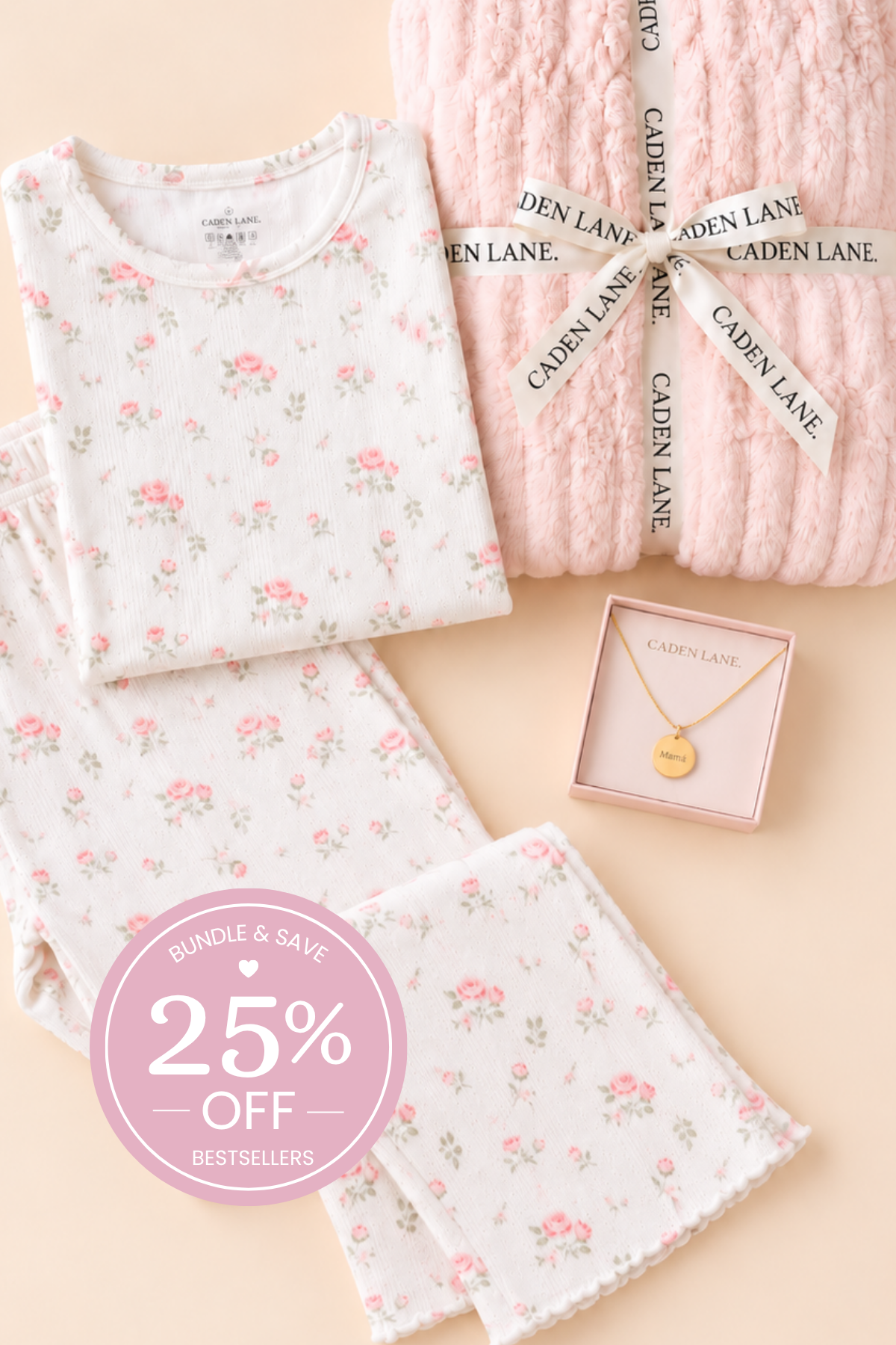 A flat-lay of Caden Lane mom ultimate gift set, featuring a white long-sleeve top and matching pants with a pink and green floral print, next to a chunky light pink knit blanket tied with a branded ribbon, and a small gold pendant necklace in a pink box. A pink circular badge in the corner advertises a 25% off bundle.