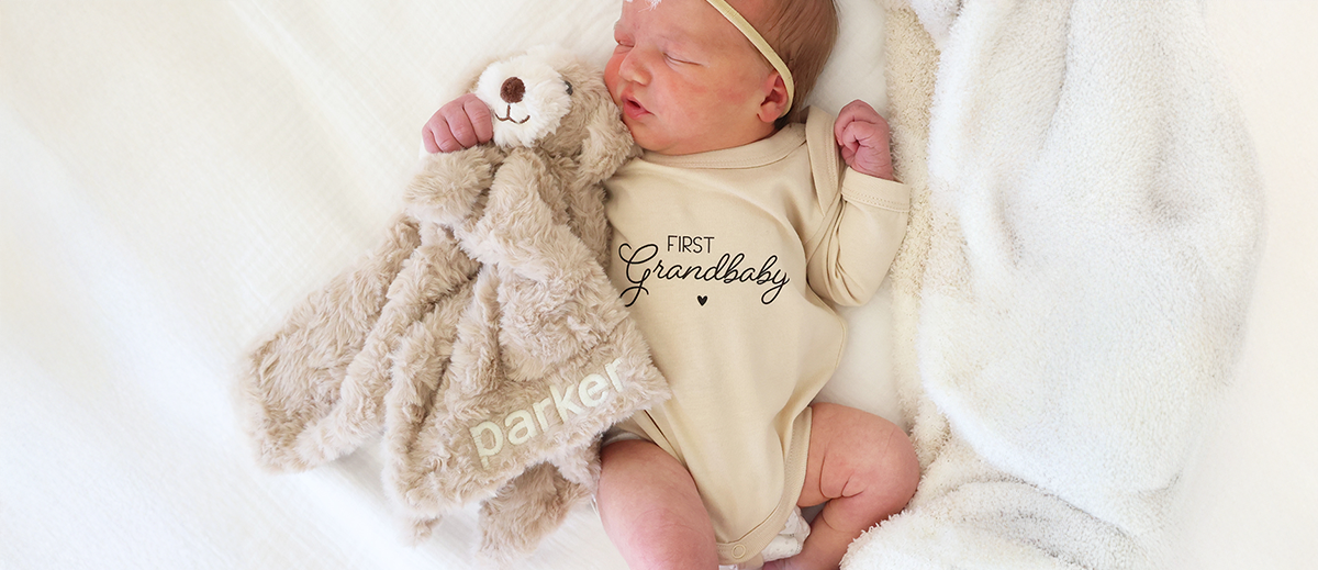 Baby wearing a first grandbaby graphic bodysuit with a personalized animal lovey from Caden Lane