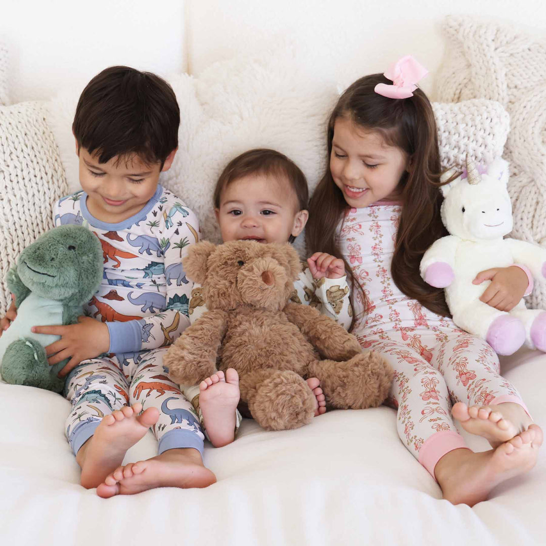 Boy kid on the left wearing a dino 2 pc pajama set and holding a trex stuffed animal. Baby in the middle holding the bear stuffed animal. Girl on the right wearing the ballet blossoms 2 pc and holding a unicorn.