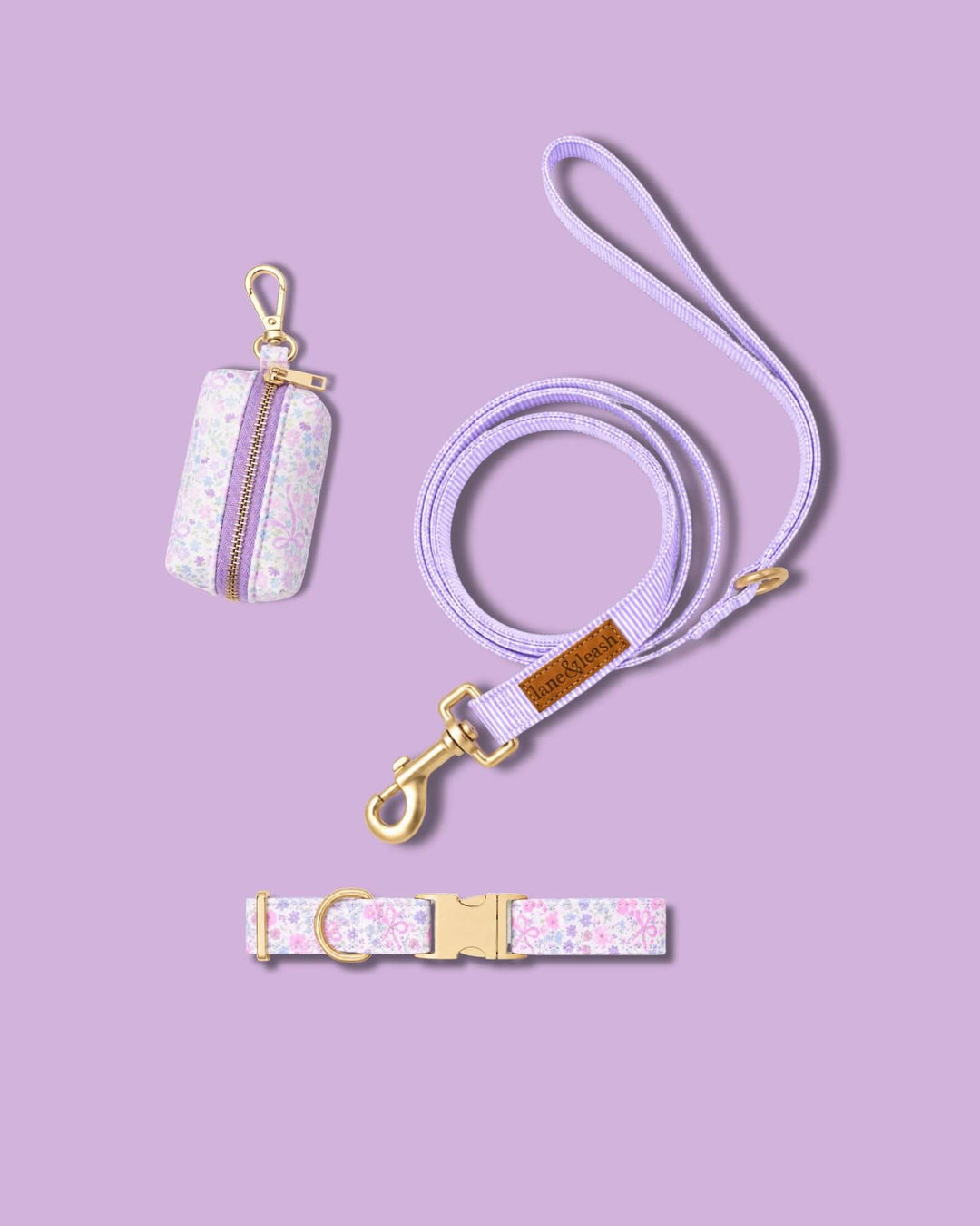 Ribbon Garden collection accessories flat lay on a lavender background: waste bag holder in pink floral print, matching purple leash, and pink floral print dog collar with gold hardware