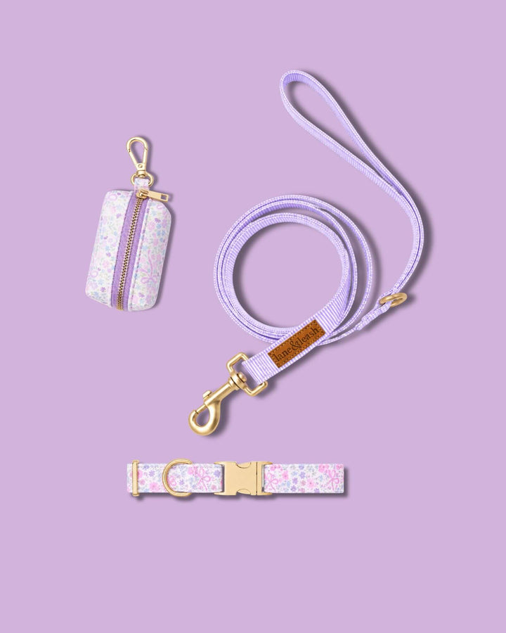 Ribbon Garden collection accessories flat lay on a lavender background: waste bag holder in pink floral print, matching purple leash, and pink floral print dog collar with gold hardware