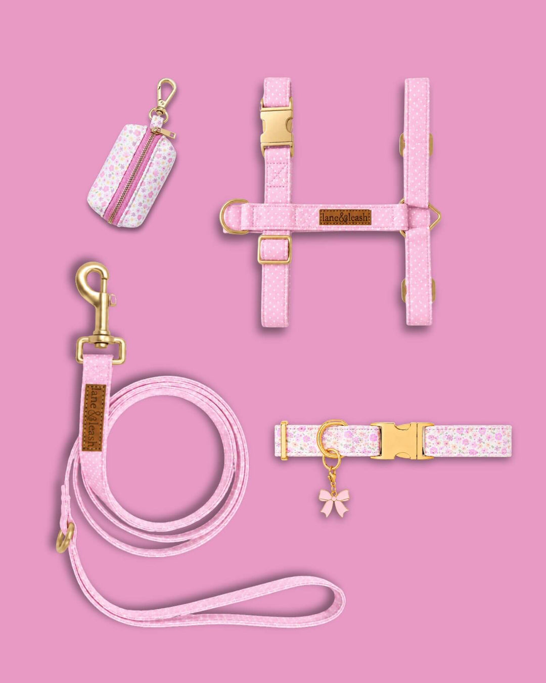 Pink floral pet accessories flatlay on pink background showing matching harness, leash, collar with pink bow charm, and poop bag holder