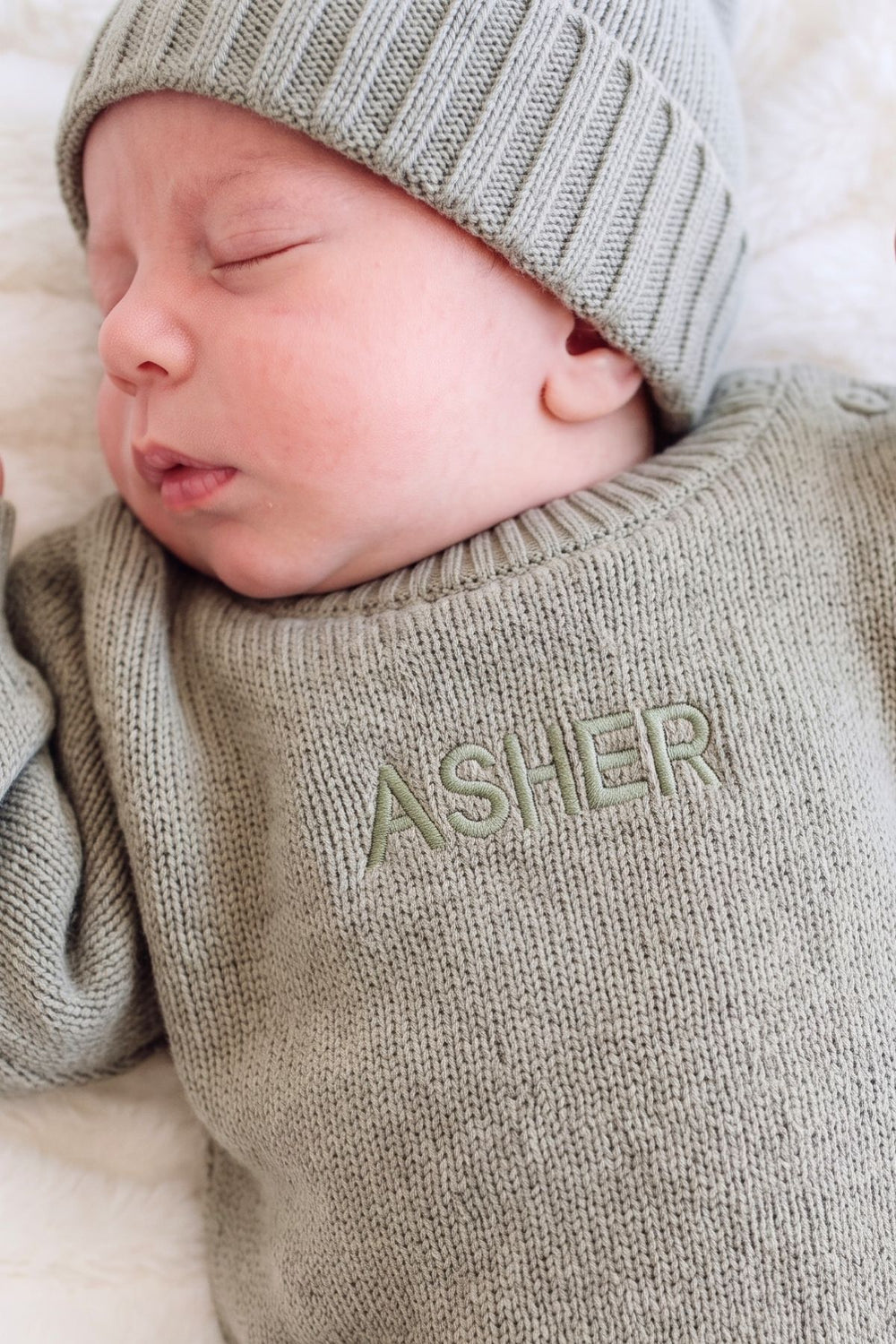 A close-up of a sleeping newborn baby wearing a Caden Lane light sage green knit sweater and matching beanie, with the name 'ASHER' embroidered on the chest.