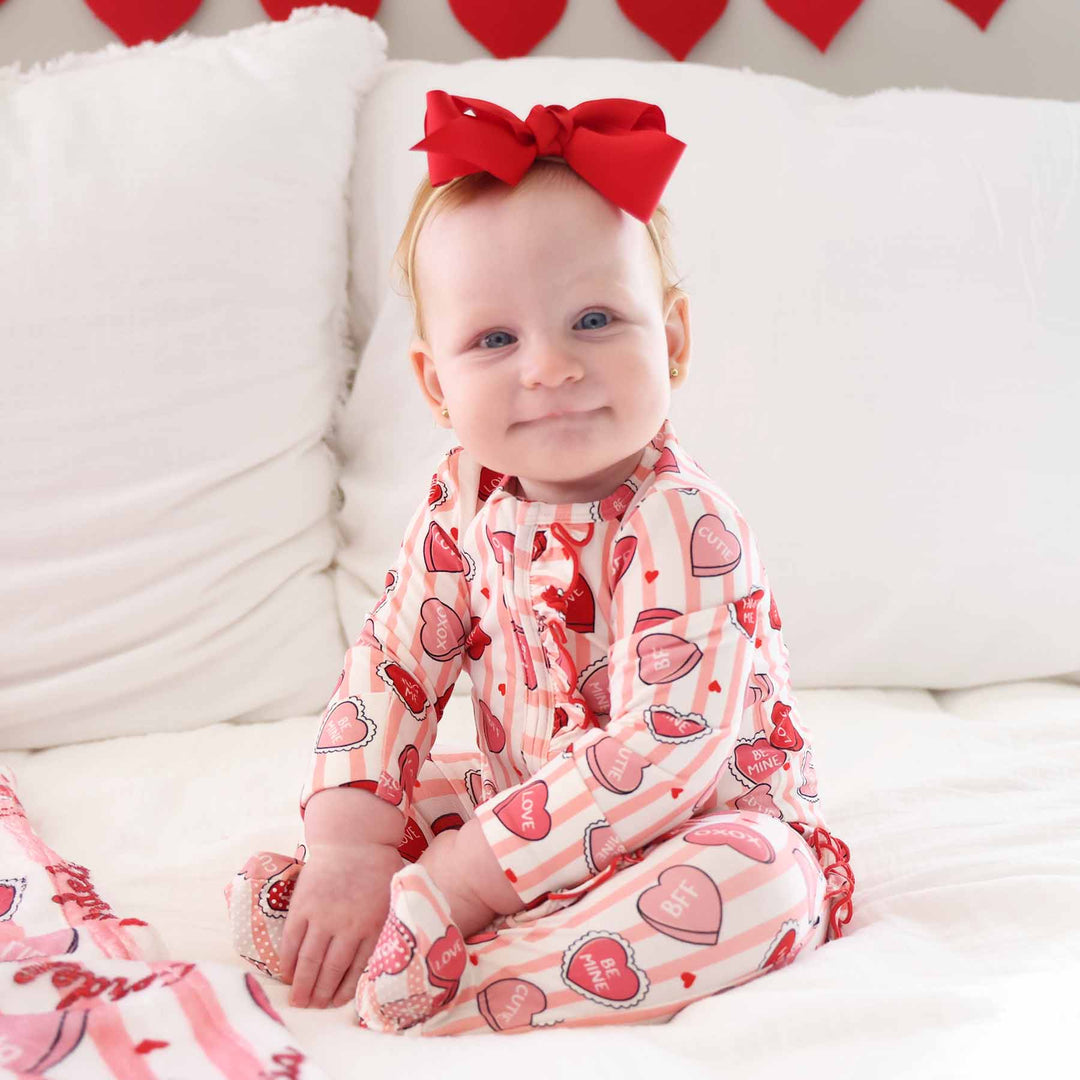 baby girl sitting on a bed in the pink sweet like candy ruffle zipper footie with a red ribbon bow on her head