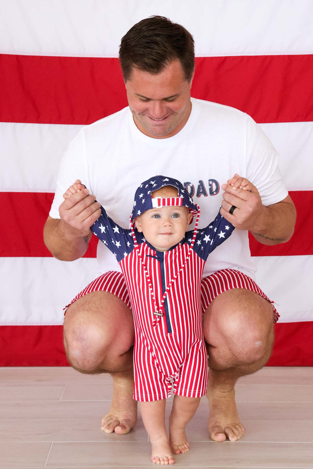 dad squatting down with baby boy in stars & stripes swimsuits 