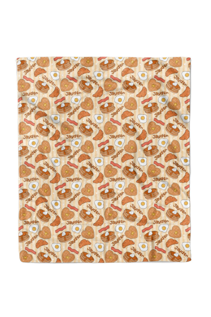 A Caden Lane blanket featuring a repeating cartoon breakfast pattern of eggs, bacon, pancakes, and toast on a light beige background.