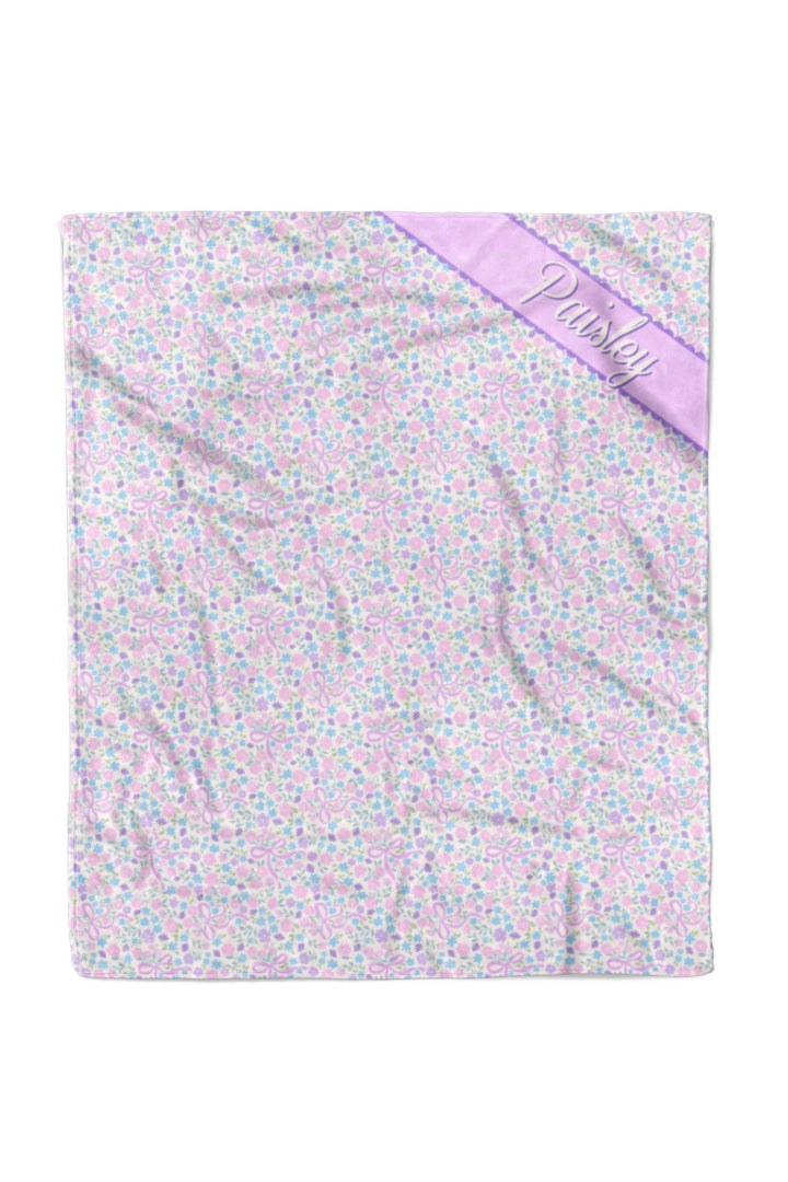 Personalized pet blanket in the Ribbon Garden print — soft pink and lavender floral pattern — with a purple satin corner ribbon personalized with the name "Paisley", shown flat on white background