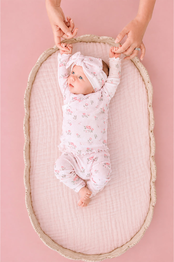 Caden Lane Rose Garden newborn take-home outfit, baby held by parent's hands in bassinet wearing pink floral footie and bow headband
