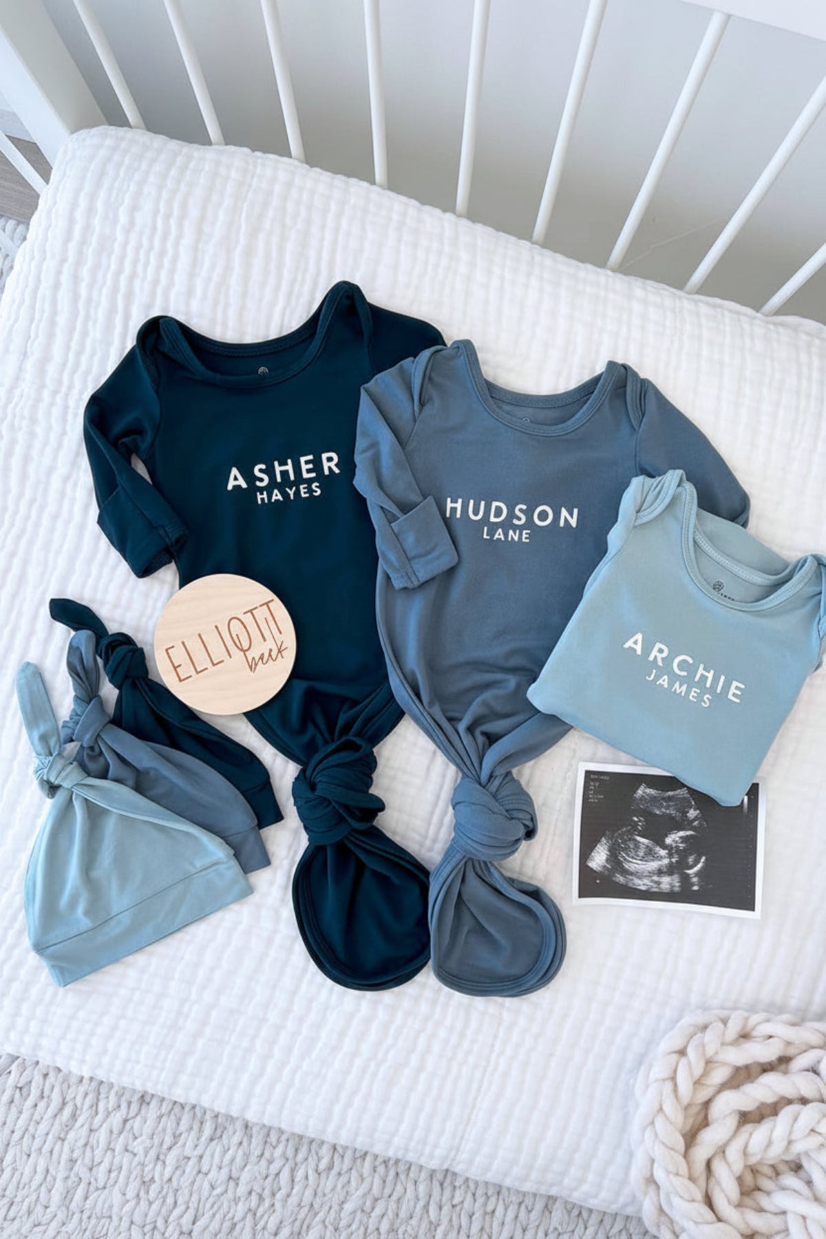 Three Caden Lane personalized knot gowns in navy, slate blue, and light blue with "Asher Hayes," "Hudson Lane," and "Archie James" in block lettering, styled with matching bows and a wooden name sign.