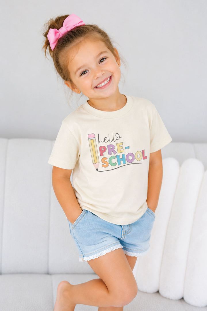 A young girl smiling and posing in a cream-colored Caden Lane t-shirt with a colorful 'hello PRE-SCHOOL' graphic, paired with light blue denim shorts.