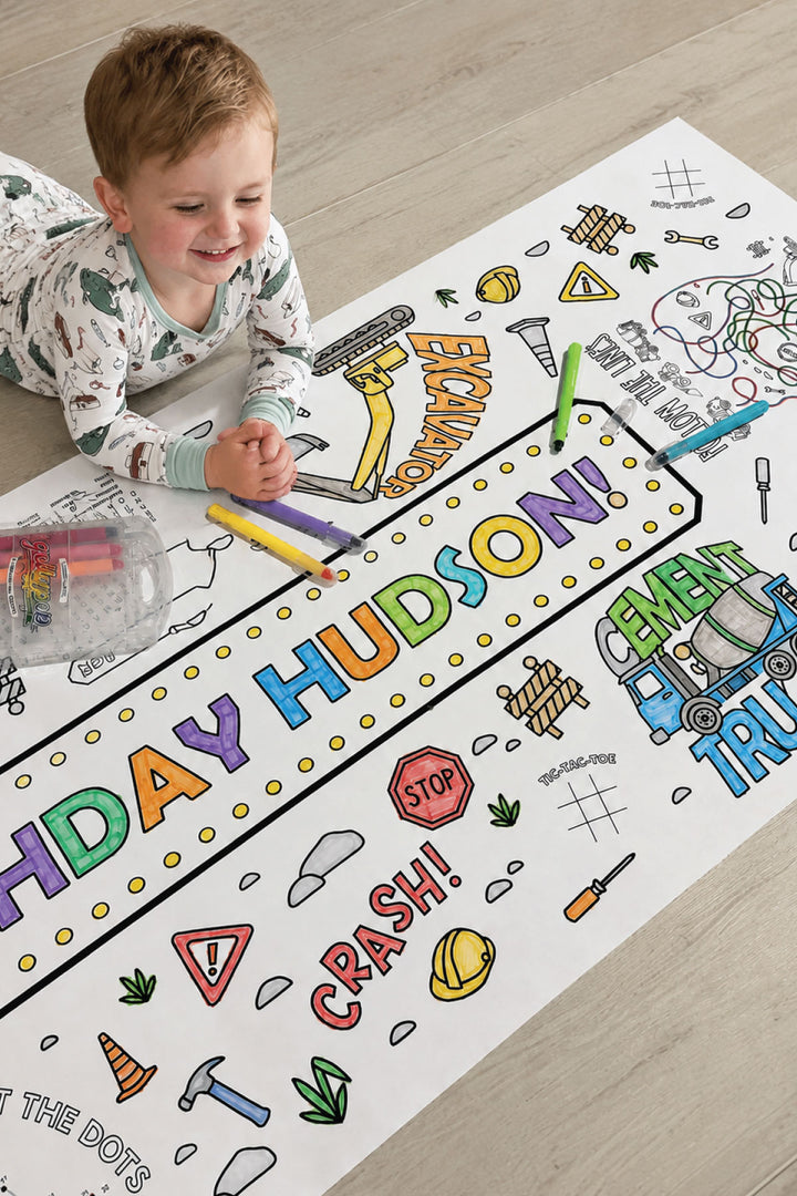 Toddler boy coloring a large construction-themed birthday banner reading 'Happy Birthday Hudson!' with colorful markers, filling in an excavator, cement truck, and stop sign illustrations.