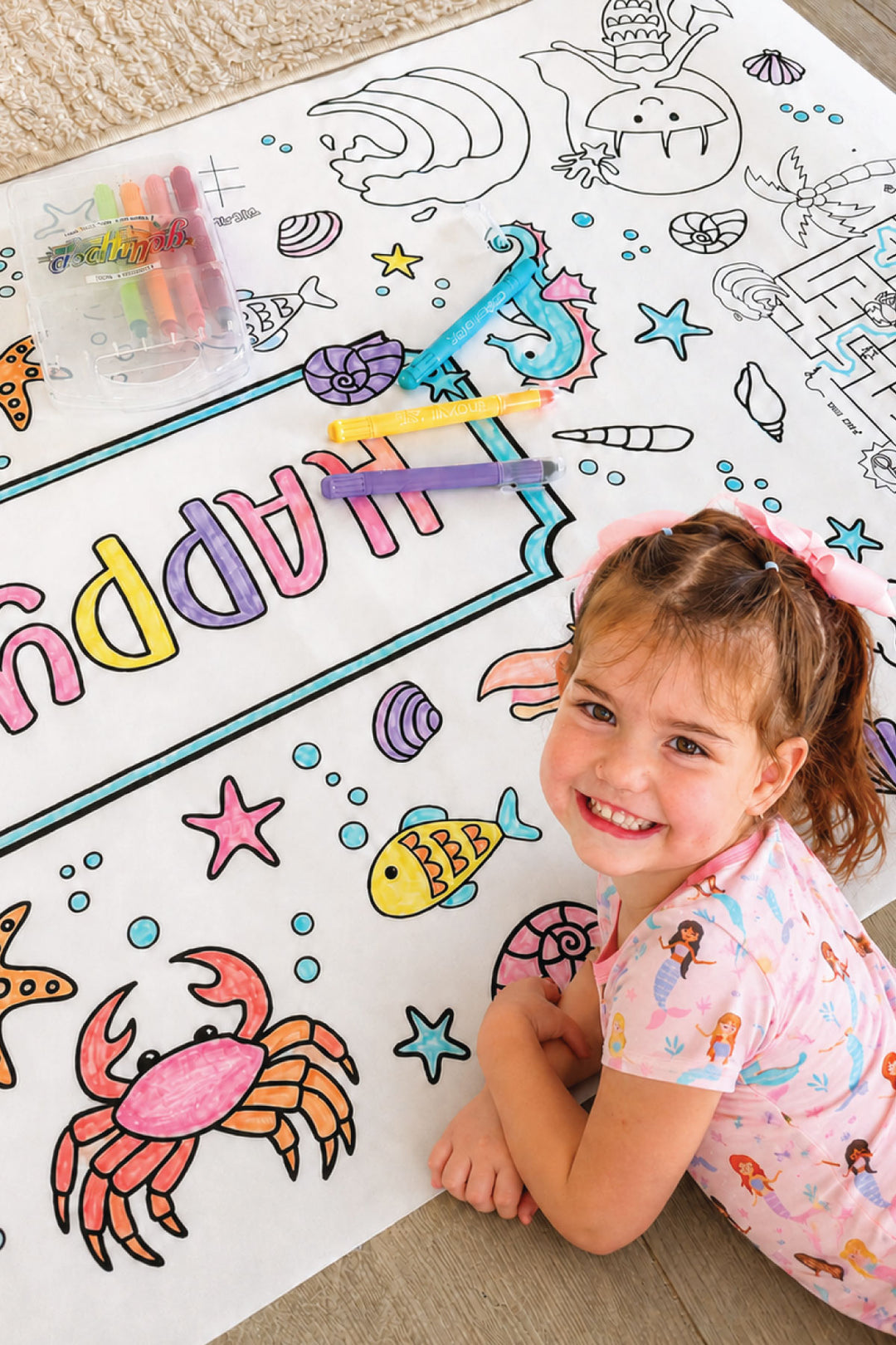 Smiling young girl in a mermaid nightgown coloring an ocean-themed birthday banner with markers, filling in a crab, fish, starfish, and seashell illustrations