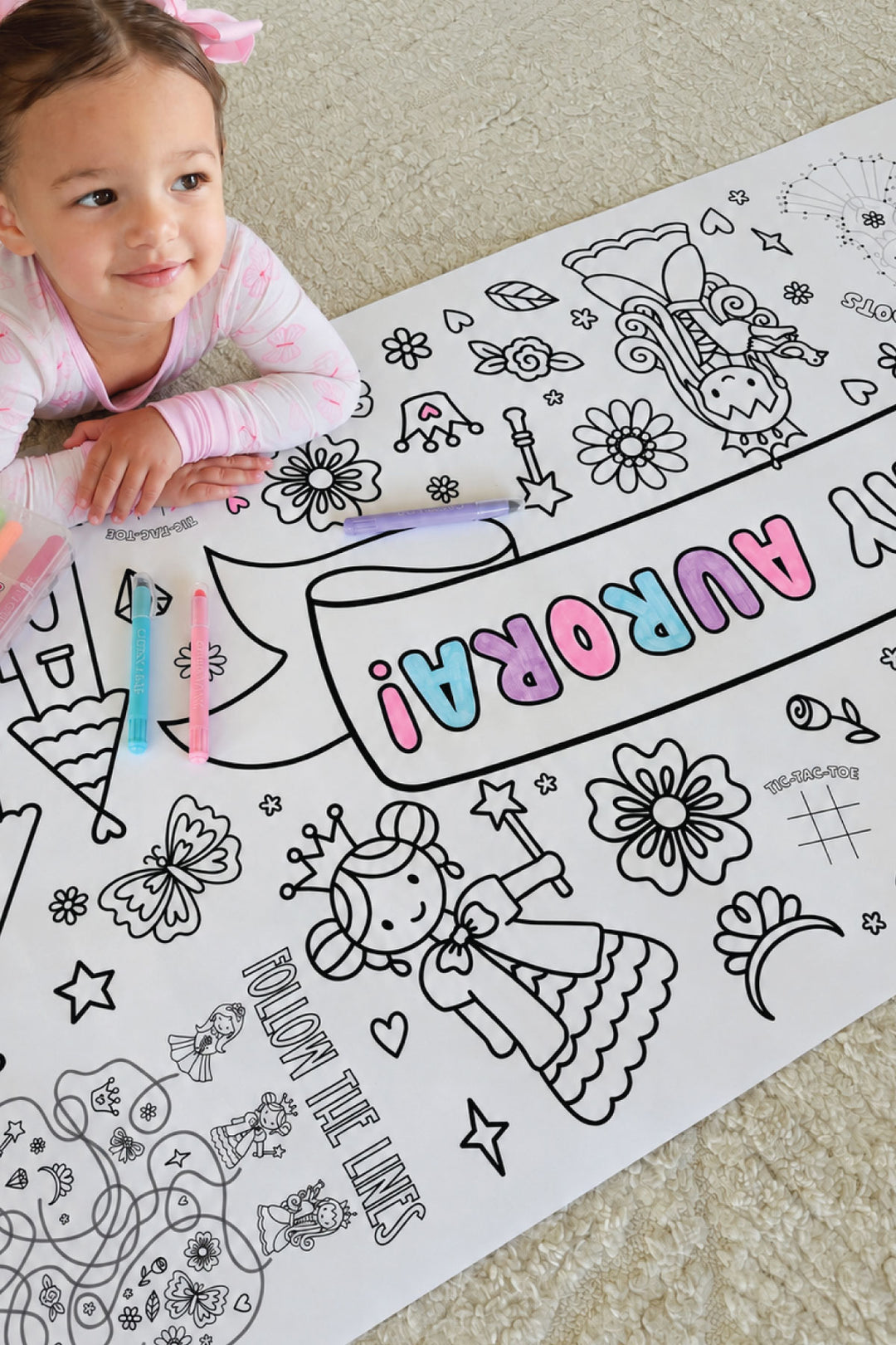 Smiling toddler girl coloring a princess-themed birthday banner reading 'Happy Birthday Aurora!' with markers, coloring in a princess, flowers, and butterfly illustrations