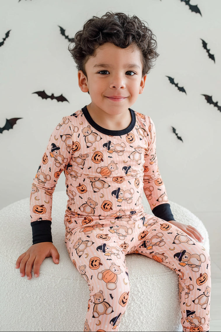 Caden Lane young boy with curly dark hair sits on a white textured stool, wearing a light orange long-sleeve pajama set with a black crew neck and cuffs, and an all-over Halloween print of pumpkins, ghosts, and bats. He is holding a small green candy in a brightly lit room with black bat decals on the wall.