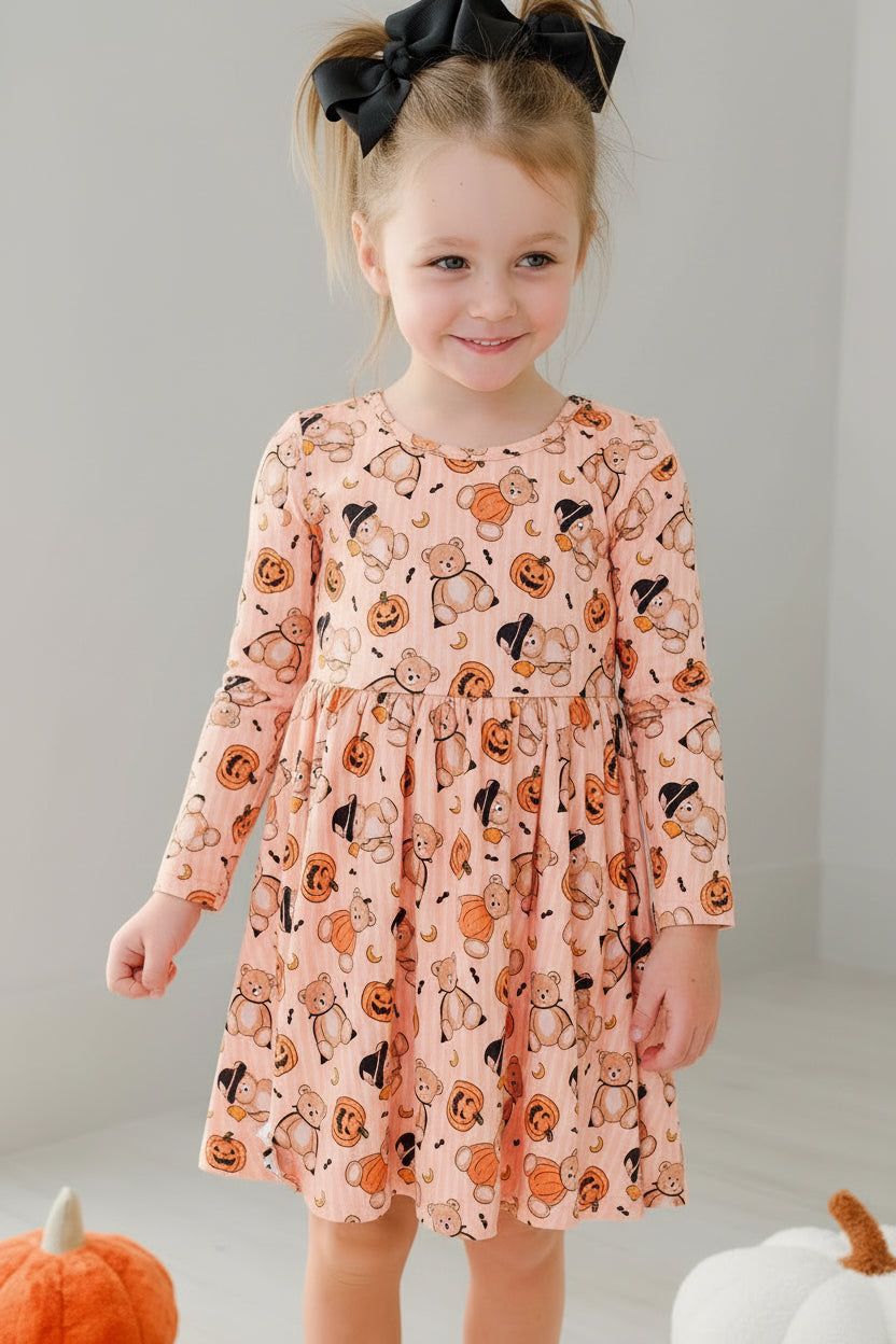 Caden Lane young girl stands in a bright room wearing a long-sleeve orange dress with an all-over black cat print. She has a large black hair bow in her hair and is posed next to decorative pumpkins and a white chair.
