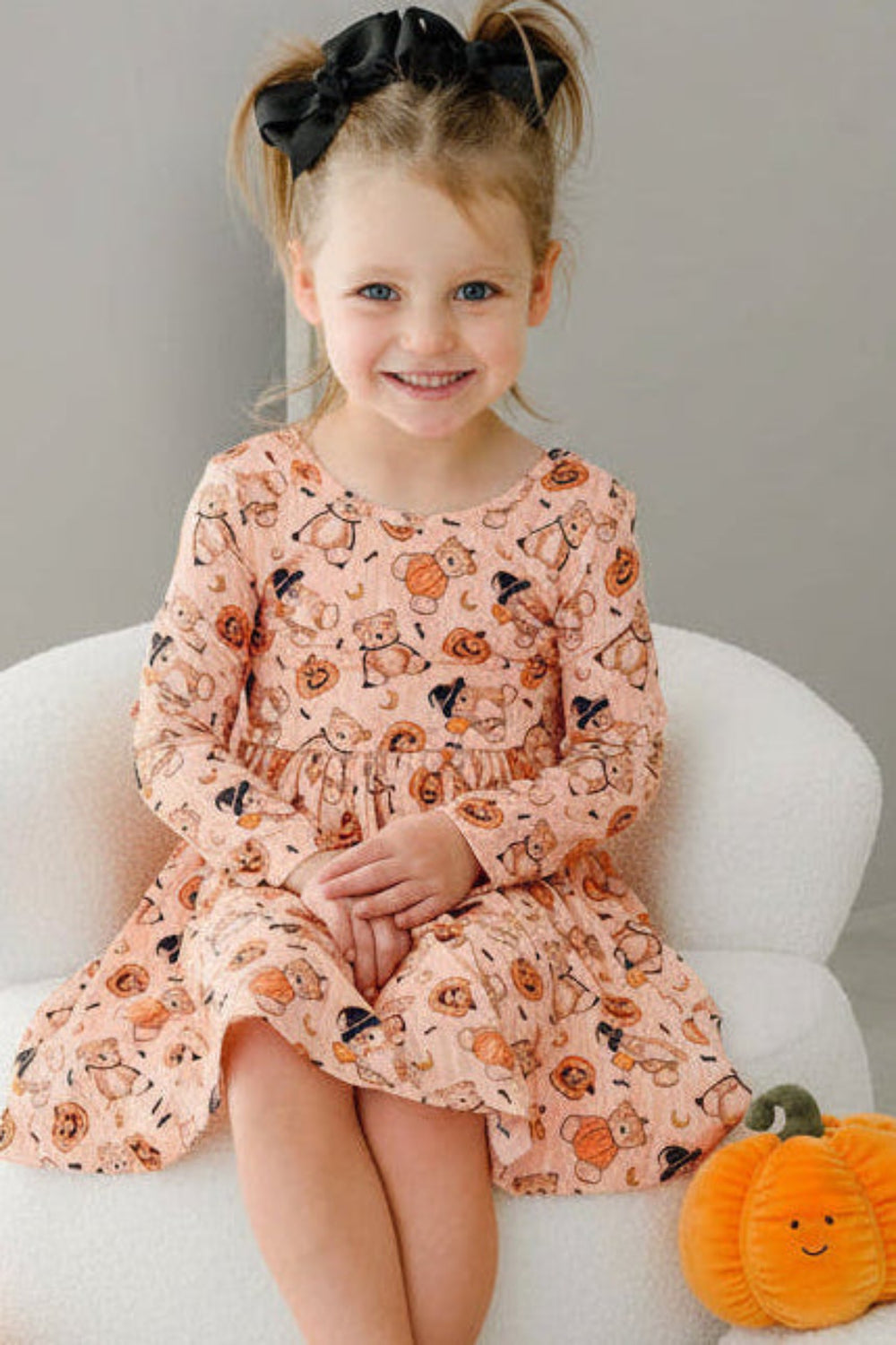 Caden Lane young girl with her hair in two buns sits on a white armchair, wearing an orange long-sleeve dress with an orange and black pumpkin print. The scene is styled with plush pumpkins on a light-colored floor against a neutral gray wall.