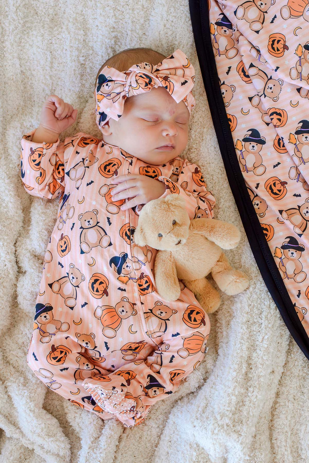 Caden Lane a sleeping newborn baby wearing an orange Halloween-themed footless sleeper and matching bow headband, lying on a cream textured blanket and holding a beige teddy bear. A matching blanket is partially visible on the right.