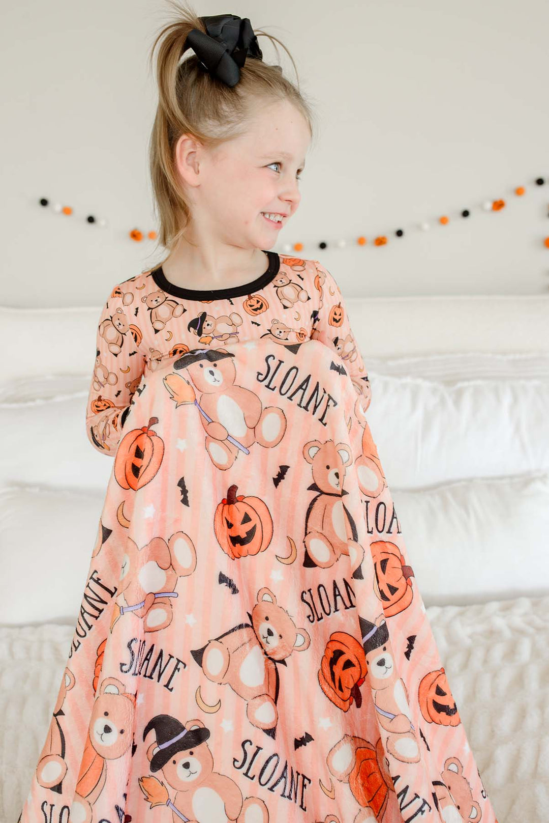 Caden Lane young girl with blonde hair in a black bow is wearing a long-sleeve halloween pajama set with a matching personalized blanket in her hands