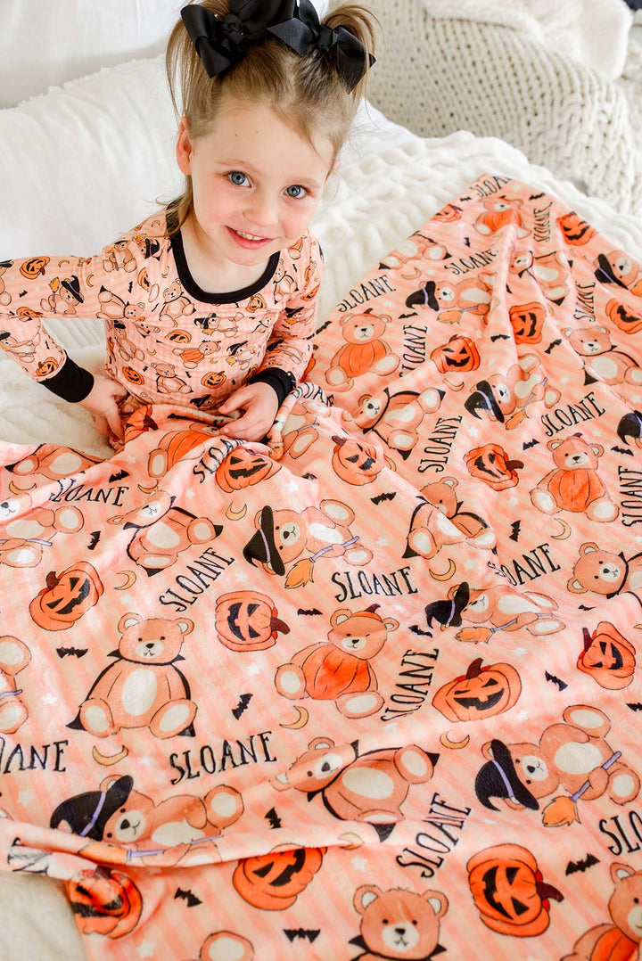 Caden Lane young girl with a black bow in her hair sits on a white bed, wearing a peach-colored pajama top and matching blanket set with a Halloween print of pumpkins, bats, ghosts, and the word 'Halloween'.