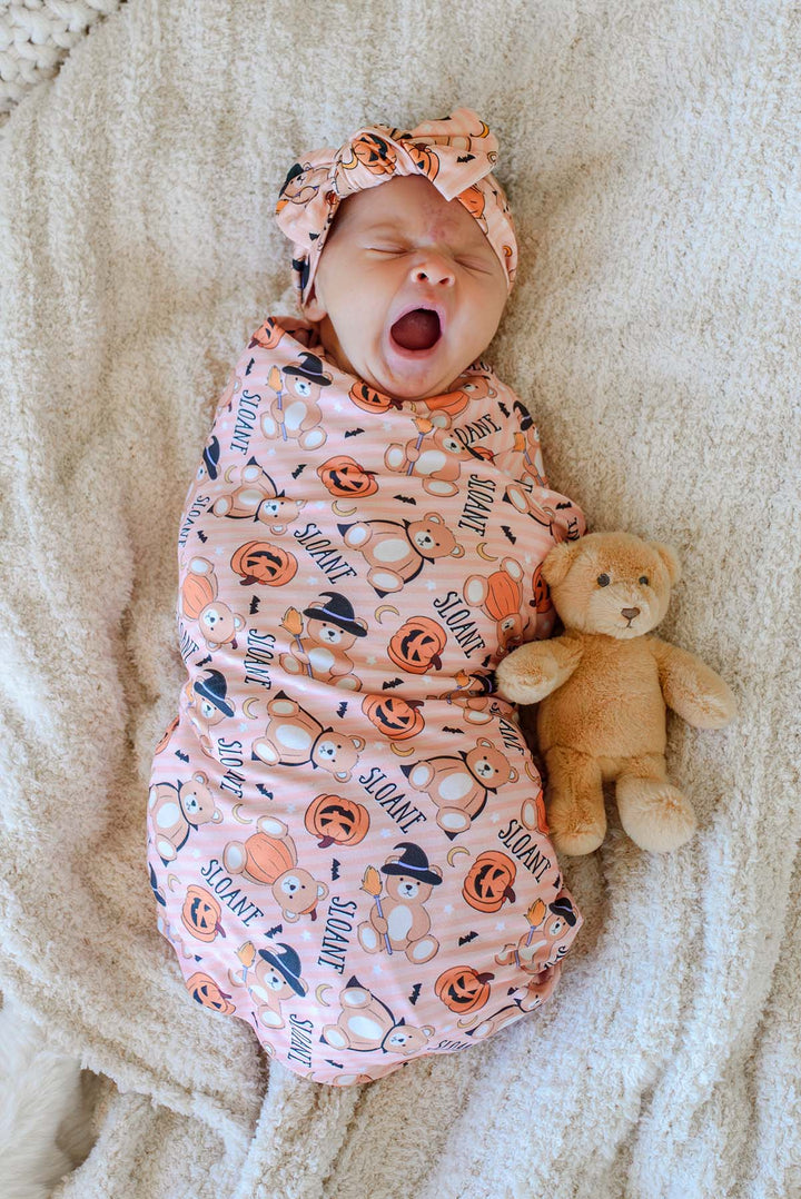 Caden Lane A newborn baby wearing an orange Halloween-themed swaddle blanket and headwrap, lying on a cream blanket next to a teddy bear.