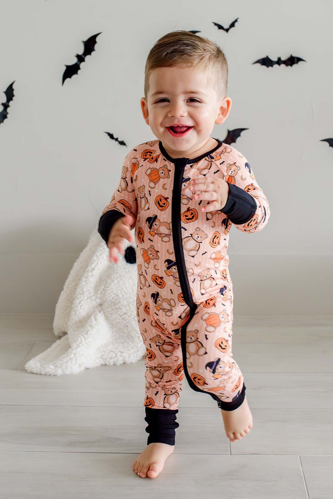 A toddler boy wearing an orange Halloween-themed zip-up sleeper with black trim and an orange pumpkin print, walking barefoot in a brightly lit room with bat decorations on the wall.