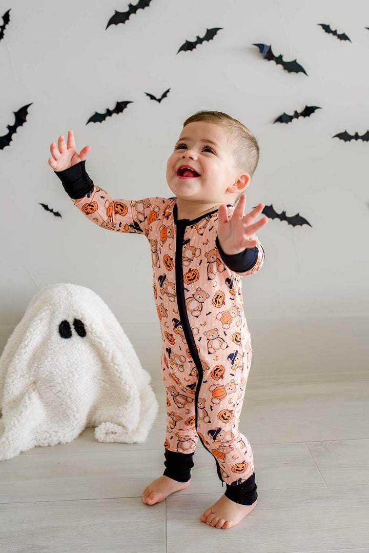 A baby standing barefoot in a Halloween-themed onesie, with a white ghost plush toy on the floor and bat decorations on the wall behind.