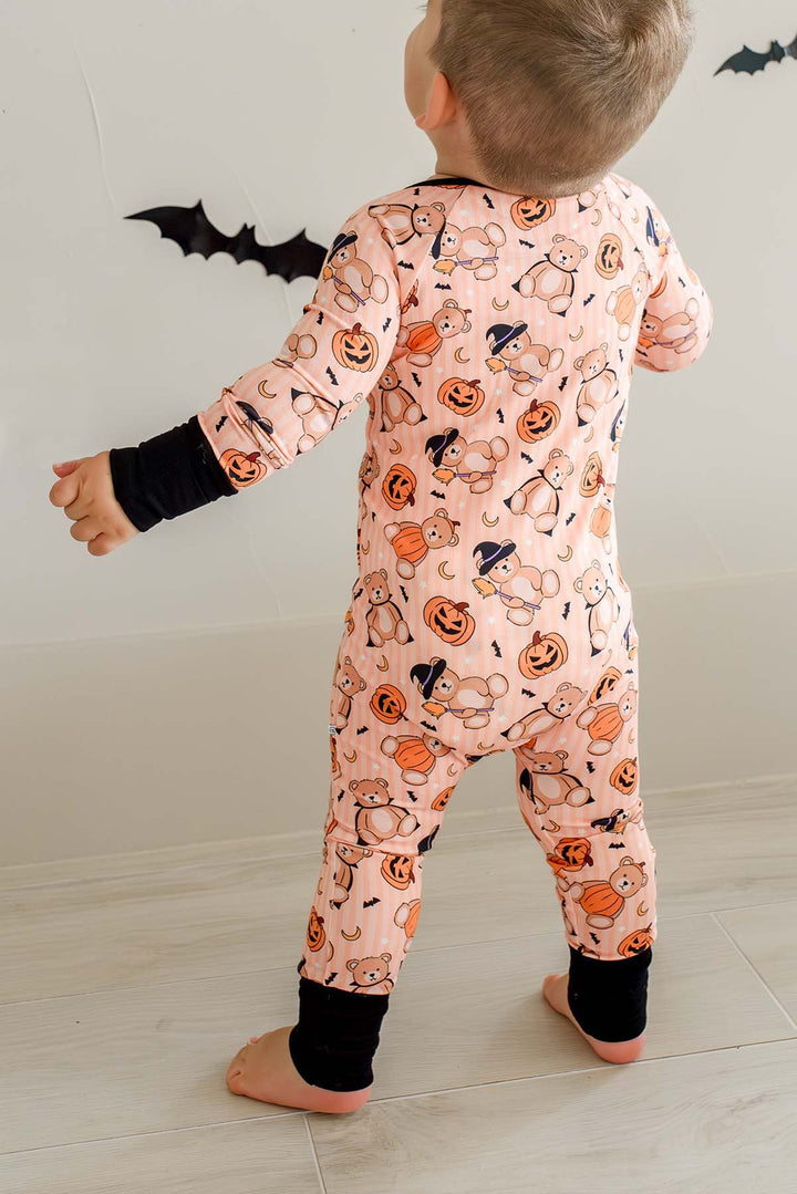 A toddler standing with their back to the camera, wearing an orange long-sleeve footed pajama romper covered in a Halloween print of pumpkins, ghosts, and bats, with black cuffs.