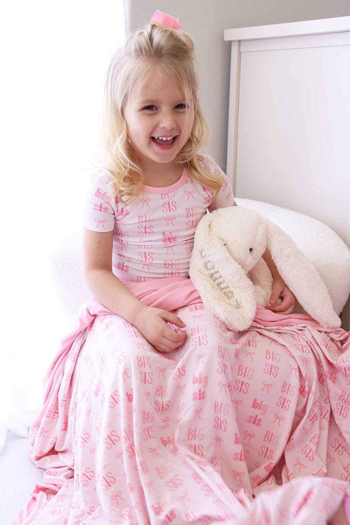 girl sitting on chair in pink two piece pajama set with bamboo blanket over her lap and holding an embroidered bunny stuffed animal 