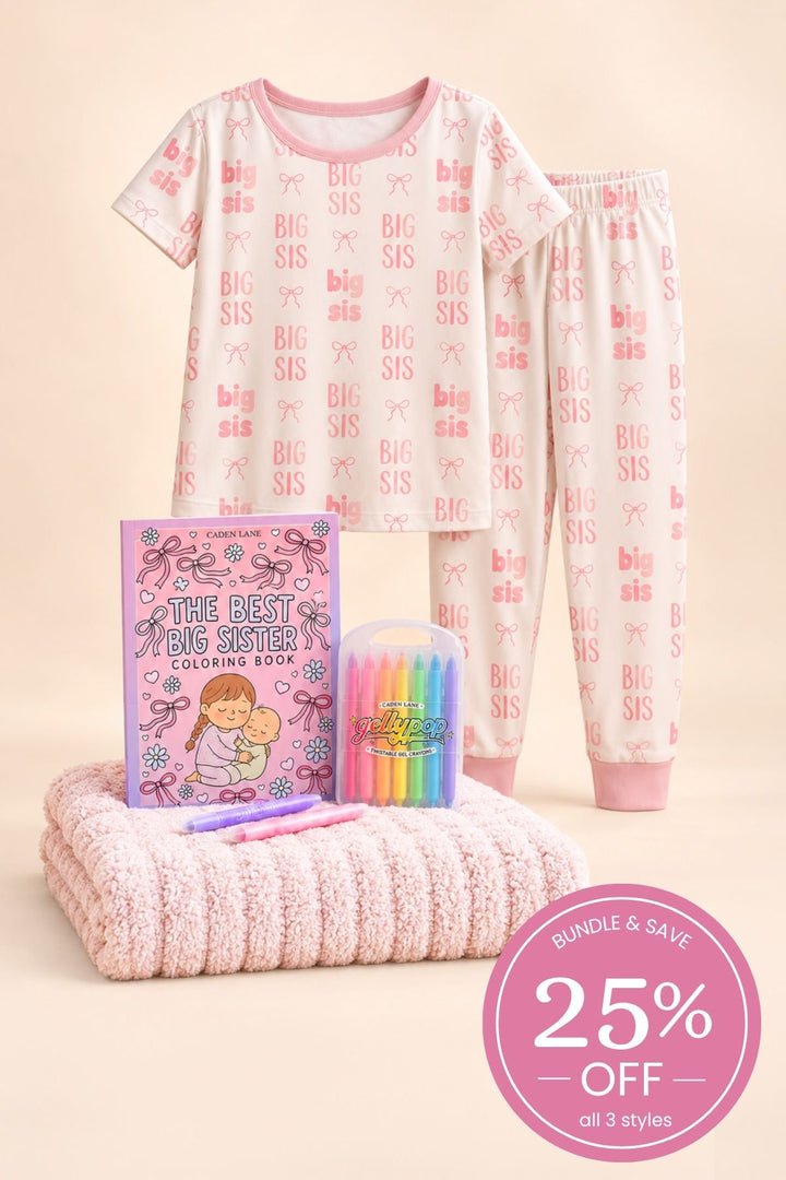 Product shot of a Caden Lane pink bamboo short-sleeve pajama set in a Big Sis print featuring pink lettering and bows, paired with a coloring book, crayons and rose blanket.