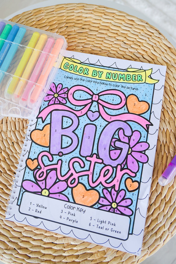 Personalized 'Camila' Big Sister color-by-number page from a Caden Lane coloring book, fully colored with a floral and bow design, displayed with a case of pastel markers