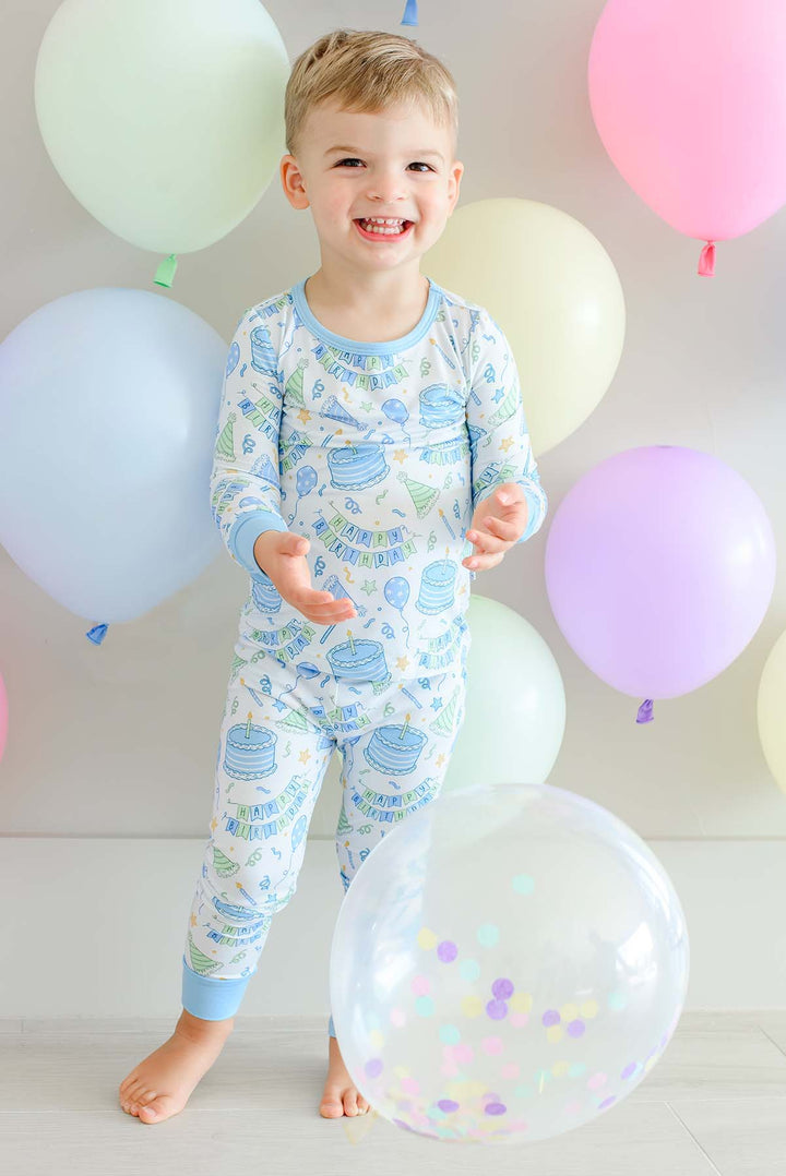 Smiling toddler boy wearing Caden Lane Birthday Bash Blue two-piece pajamas with birthday cakes, balloons and party hats.