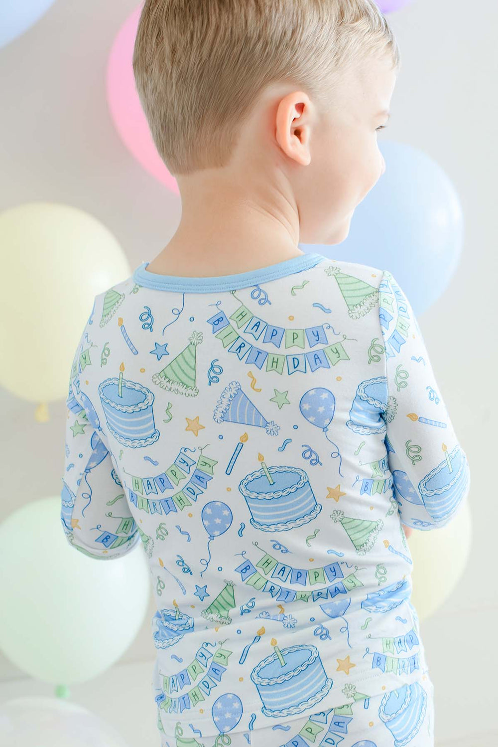 Back view of a toddler wearing Caden Lane Birthday Bash Blue two-piece pajamas with a colorful birthday party print.