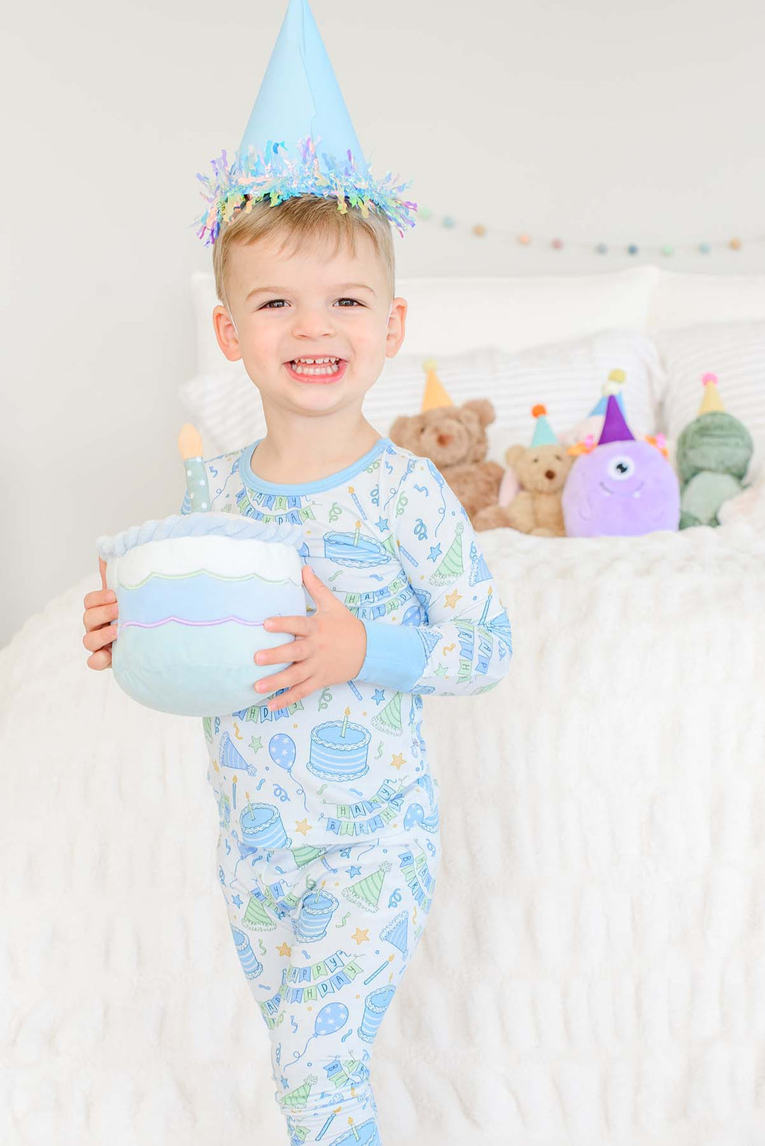 Smiling toddler boy wearing a Caden Lane Birthday Bash two-piece pajama set in blue with cake and party hat print, sporting a blue party hat and holding a plush birthday cake toy, standing by a bed with party-hatted stuffed animals.