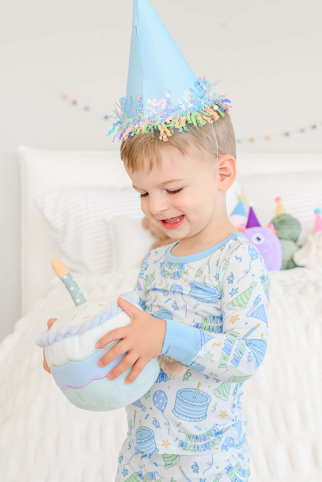 Close-up of a toddler in Caden Lane Birthday Bash Blue kids’ pajamas holding a plush birthday cake and wearing a party hat.