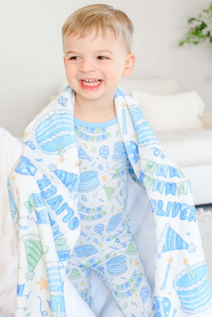 A smiling young boy wrapped in a Caden Lane white blanket with a blue and green birthday party pattern, wearing a matching light blue pajama set in a bright, modern room.