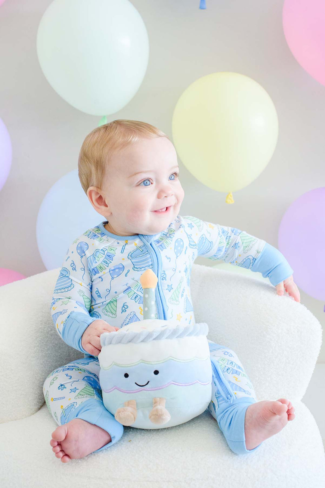 A baby sitting on a white chair wearing a Caden Lane light blue and white patterned footed pajama set, holding a matching plush toy. The background features pastel-colored balloons.