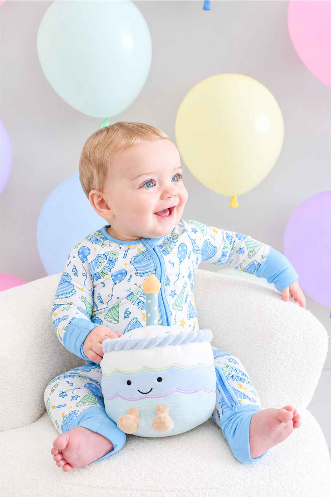 Smiling baby boy wearing a Caden Lane Birthday Bash zip footie in blue with cake and party hat print, sitting on a white chair and holding a plush birthday cake toy, with pastel balloons in the background.