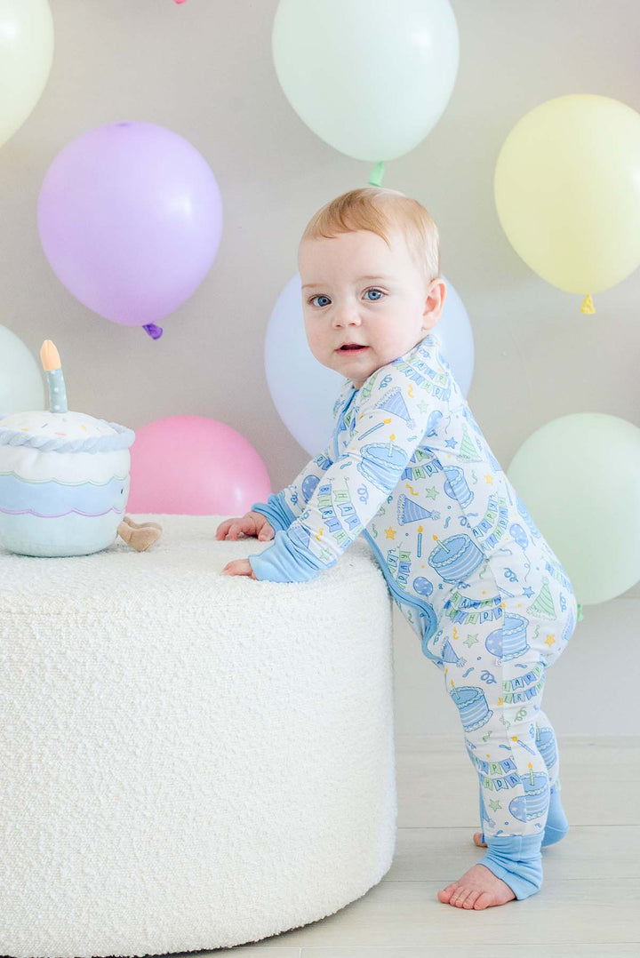 A baby standing on a white textured stool wearing a Caden Lane light blue and white patterned footed pajama, with pastel balloons in the background.