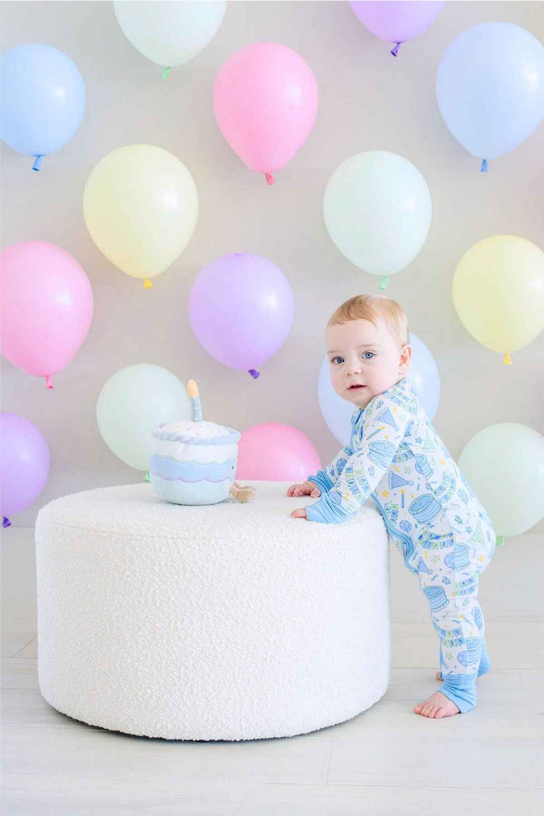 Toddler boy wearing a Caden Lane Birthday Bash zip footie in blue with cake and party print, standing beside a plush birthday cake toy atop a round white ottoman, surrounded by pastel balloons.