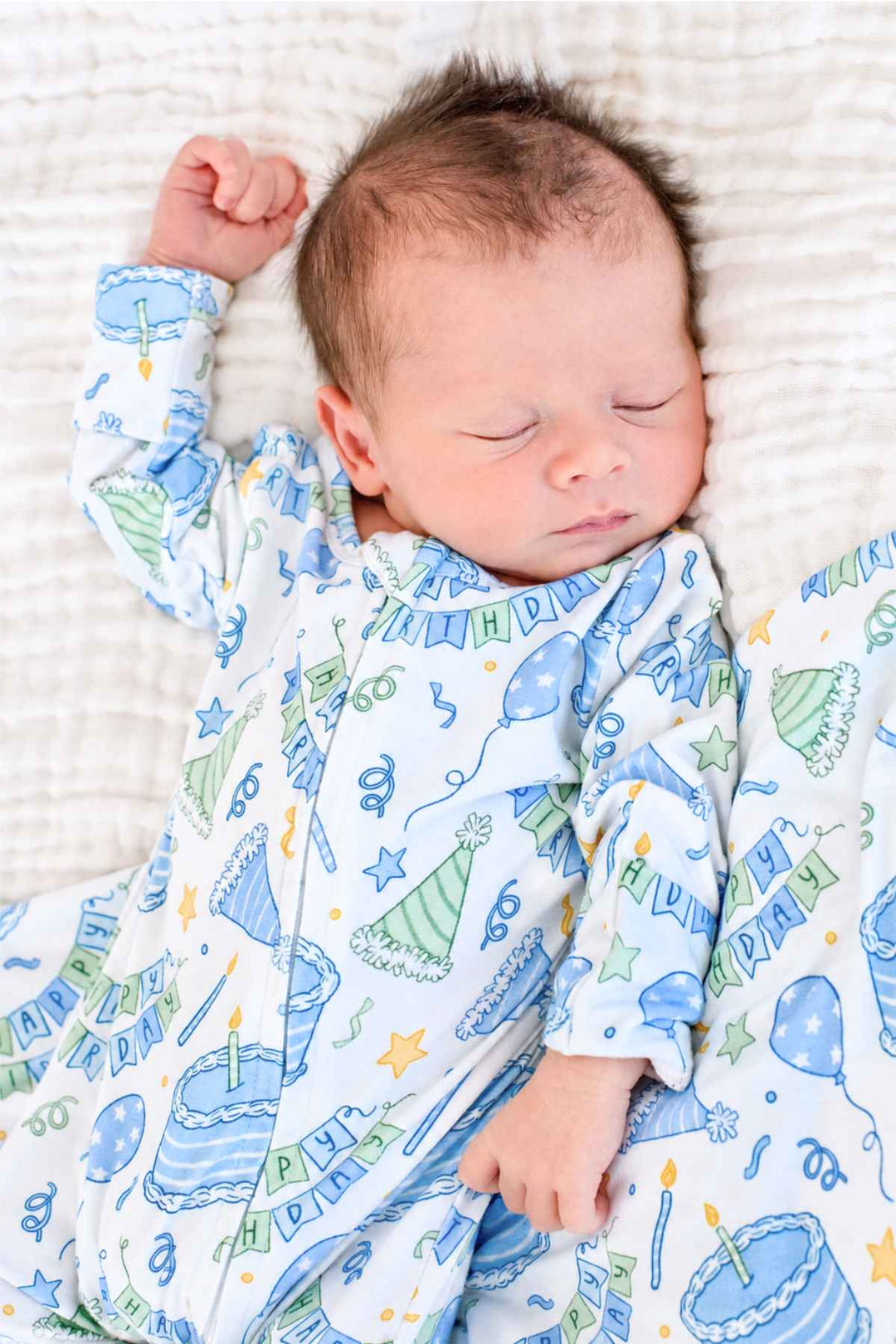 Close-up of a sleeping newborn in the Caden Lane Birthday Bash Blue baby gown with cakes, balloons and party hats.