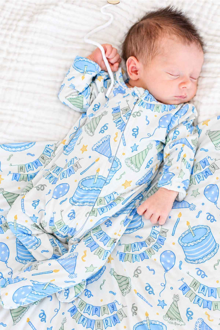 Newborn sleeping in a Caden Lane Birthday Bash Blue knotted gown beside a matching birthday-themed blanket.
