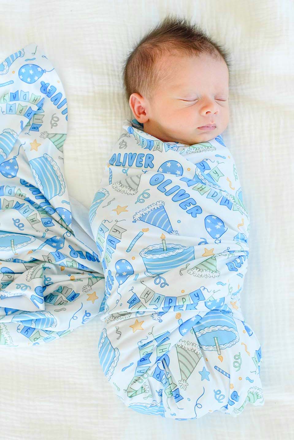 A sleeping newborn baby is swaddled in a Caden Lane personalized blue and white birthday swaddle blanket featuring the name 'Oliver', lying on a white textured blanket.