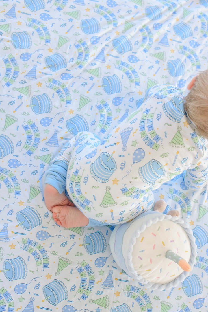 A baby wearing Caden Lane pajamas  on a matching printed blanket. The blanket has whimsical design with blue and green party hats, balloons, stars, and birthday motifs on a white background.