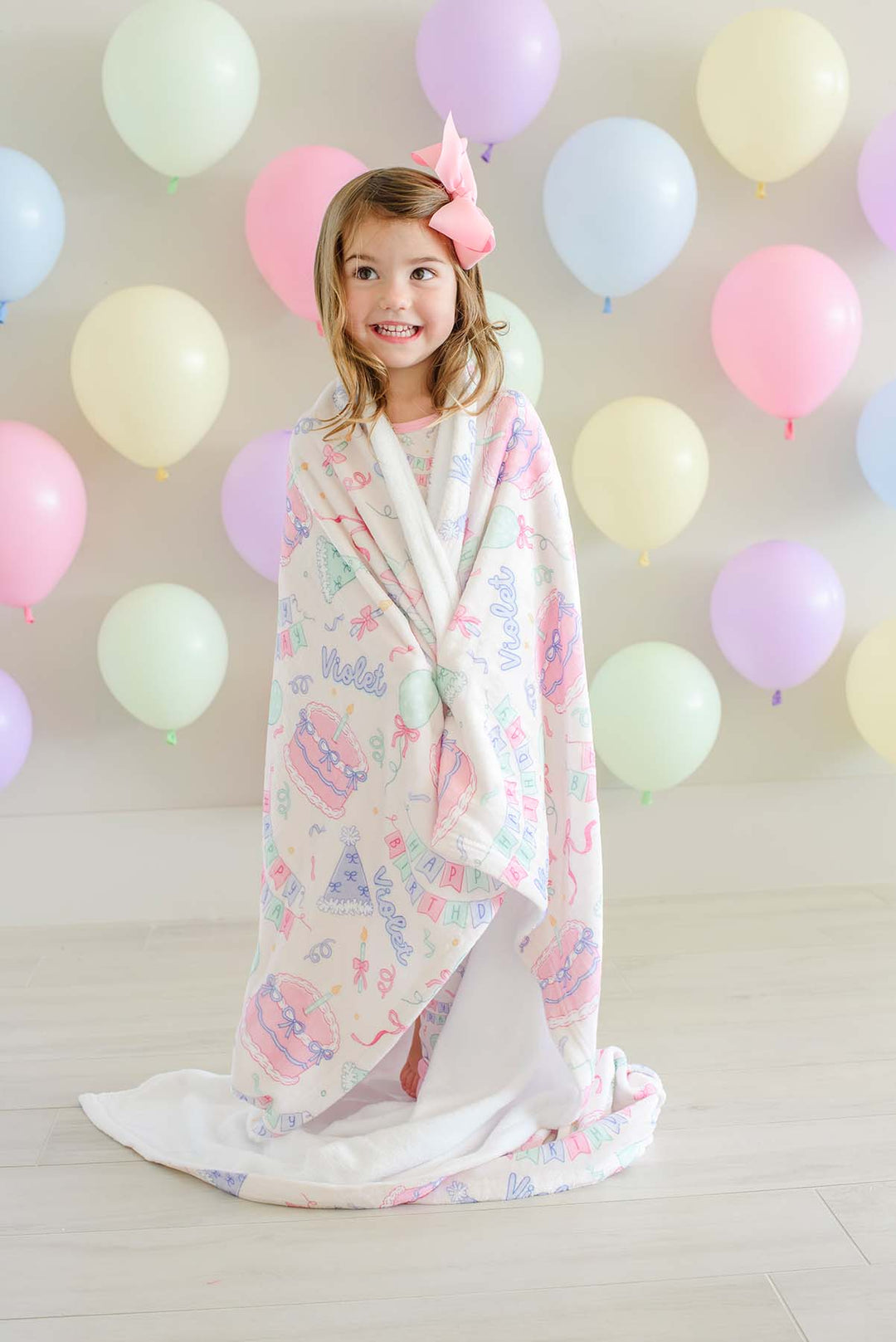 A young girl  is wrapped in a Caden Lane blanket featuring a pastel multicolor pattern of cupcakes, smiling in front of a wall decorated with pastel balloons.