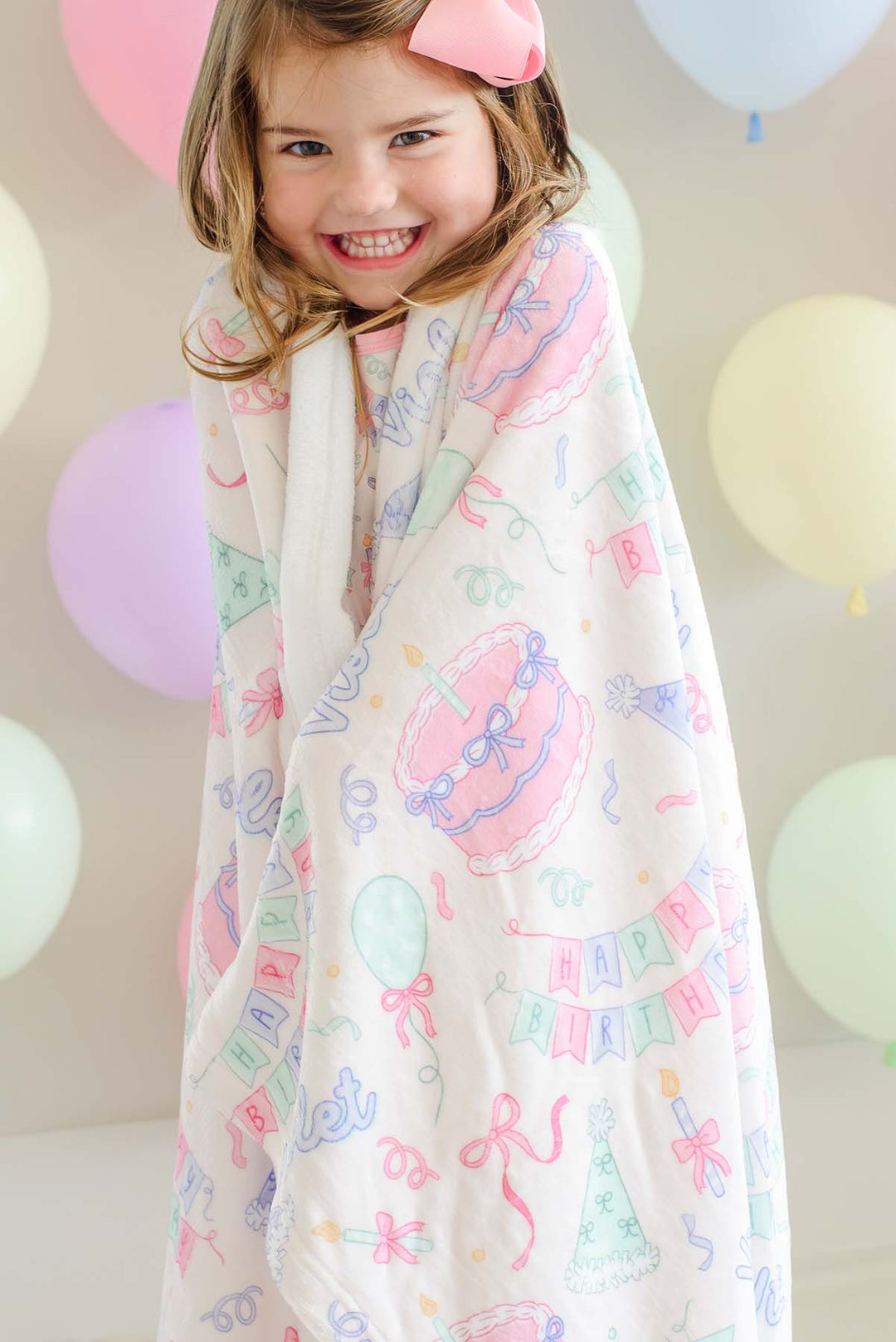 A smiling young girl wrapped in a soft Caden Lane blanket, with a pink bow in her hair. The blanket is cream-colored with a colorful pastel print of cupcakes, balloons, and 'HAPPY BIRTHDAY' banners.