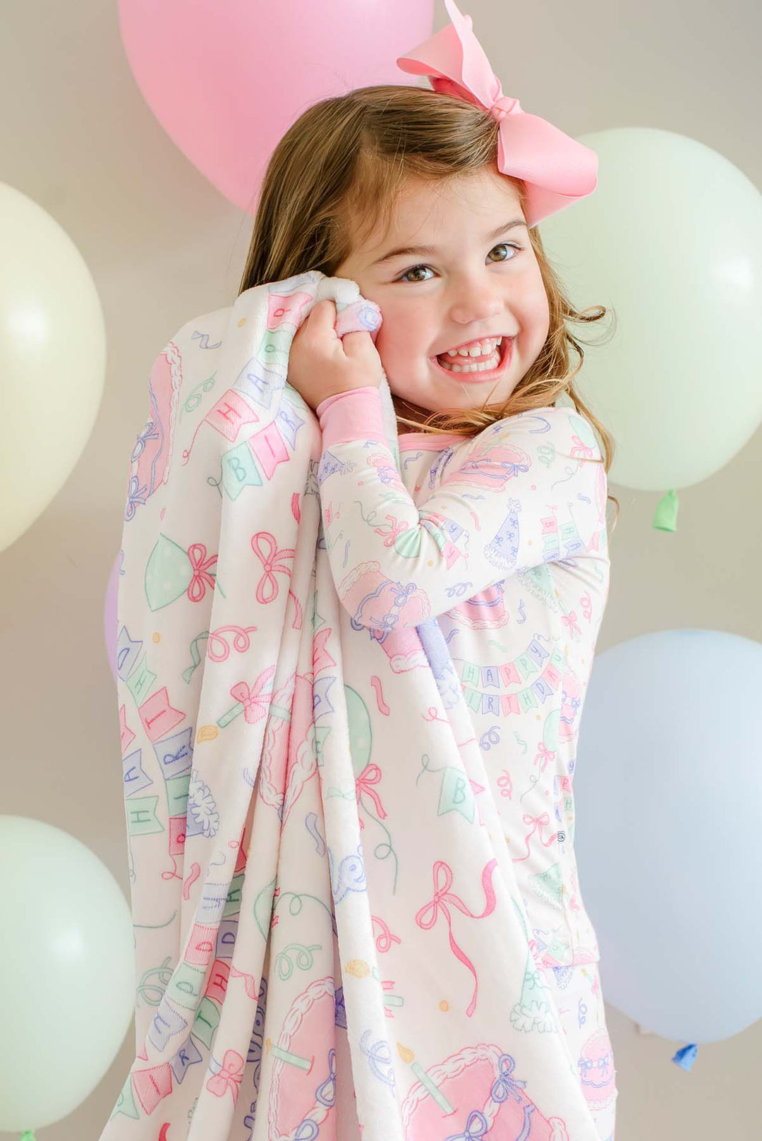 A young girl smiling and wrapping a Caden Lane pastel pink blanket with a whimsical pattern of bows, balloons, and stars. She is wearing a matching pink bow headband, set against a soft background decorated with pastel-colored balloons.
