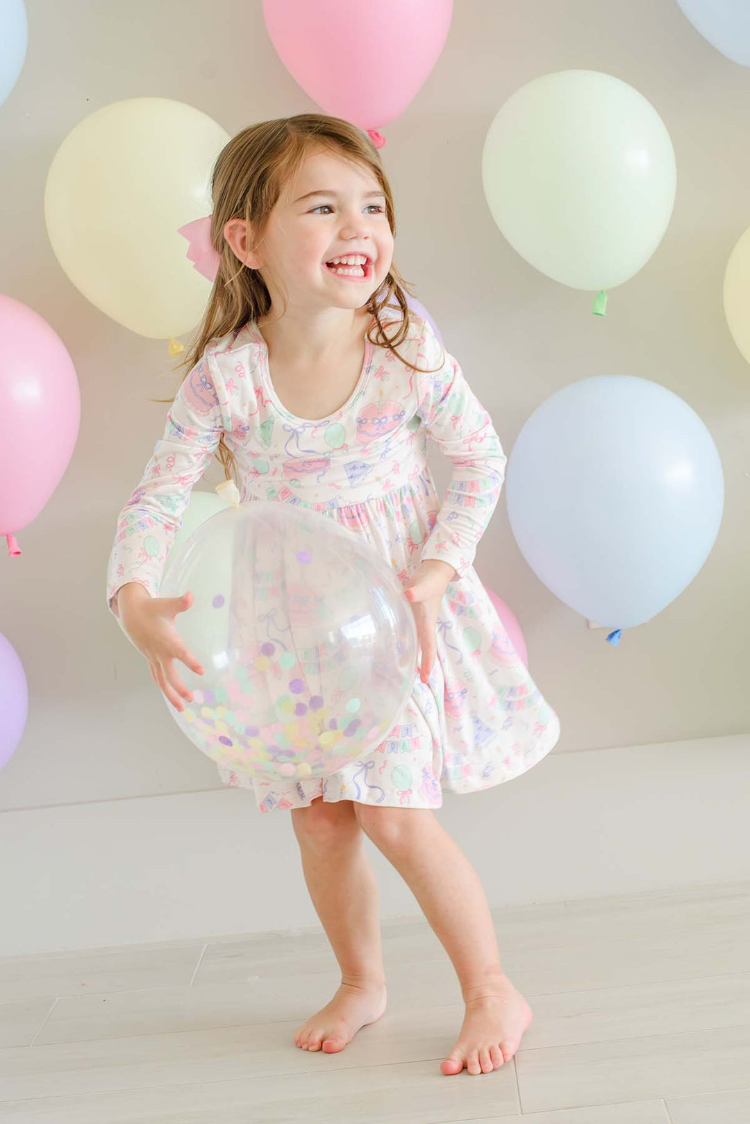 A young girl with light brown hair smiling and laughing while holding a large clear balloon. She is wearing a Caden Lane long-sleeve, knee-length dress with a white base and pastel multicolor confetti pattern.
