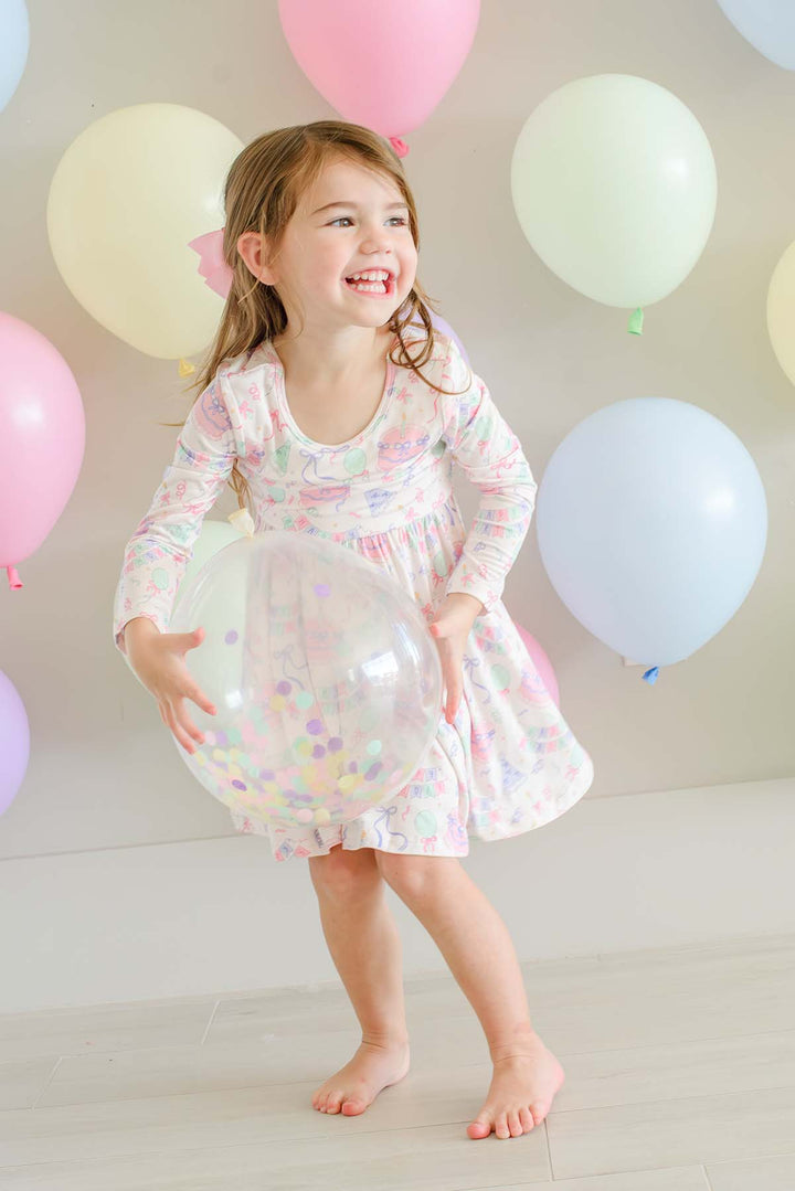 A young girl with light brown hair smiling and laughing while holding a large clear balloon. She is wearing a Caden Lane long-sleeve, knee-length dress with a white base and pastel multicolor confetti pattern.